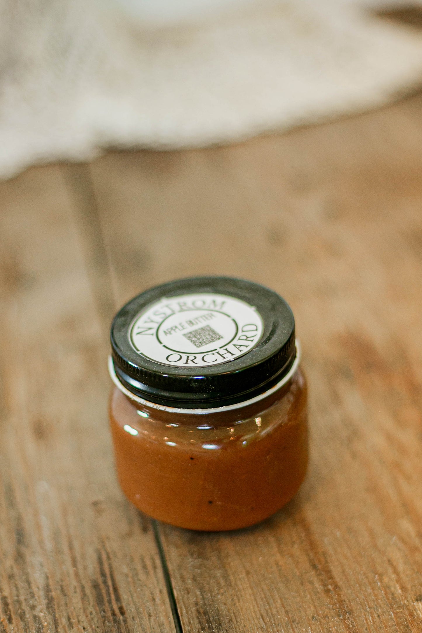 Jar of Apple Butter with a black lid on a wooden surface. Made in the kitchen of Nystrom Orchard in Worthington Minnesota.