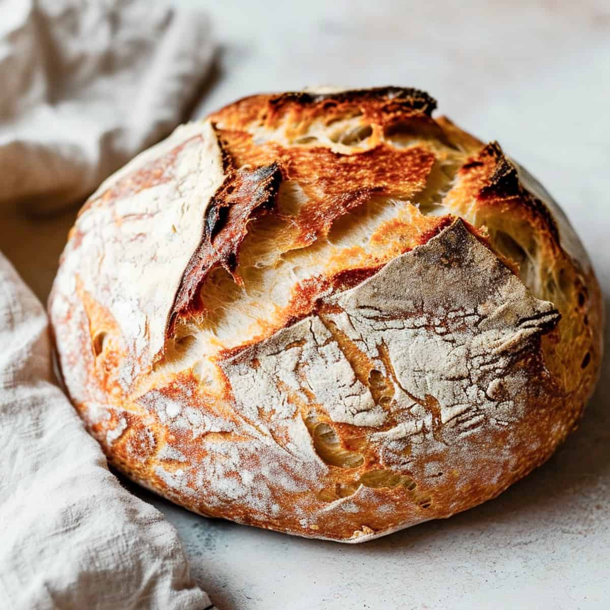 A round artisanal sourdough bread loaf with a floured crust and dark toasted spots, photographed on a light surface with a folded white cloth beside it. Baked in the kitchen of Nystrom Orchard in Worthington Minnesota.
