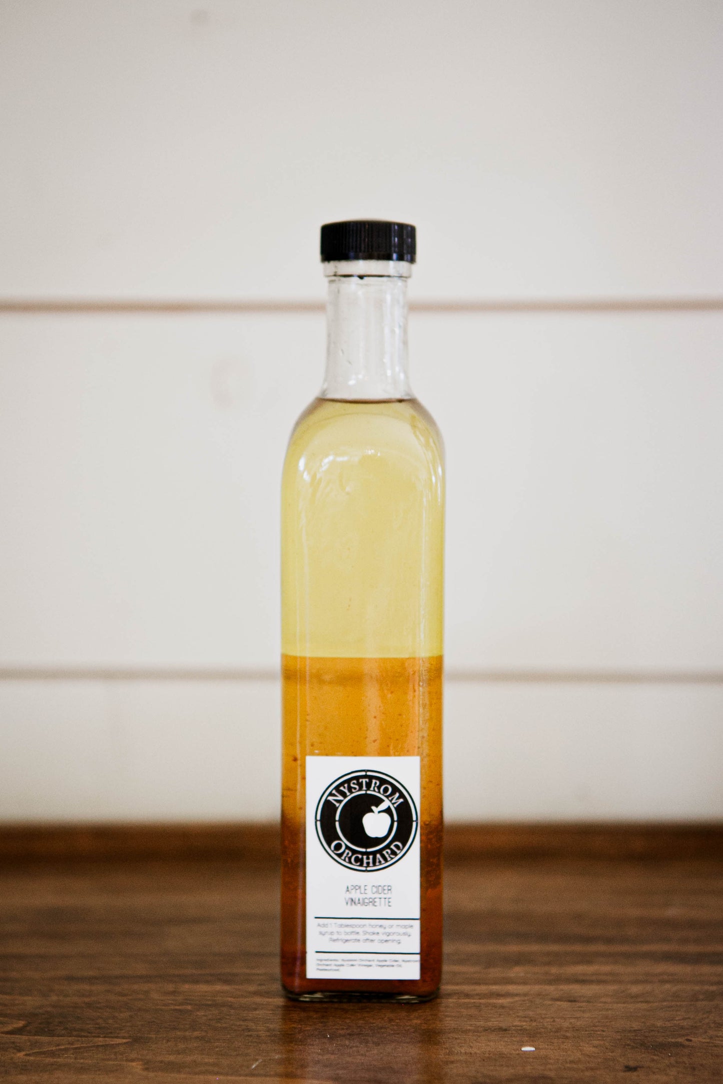 Bottle of apple cider vinaigrette made with vinegar from Nystrom Orchard on a wooden surface with a white background. Made in the kitchen at Nystrom Orchard in Worthington Minnesota.