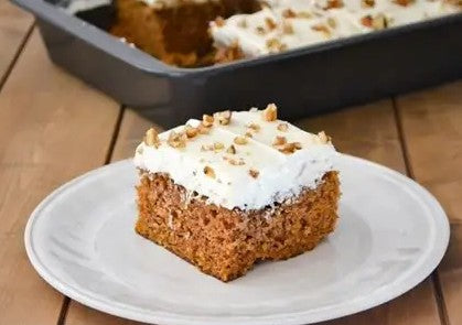 Slice of carrot cake with cream cheese frosting on a white plate, with a baking tray in the background.