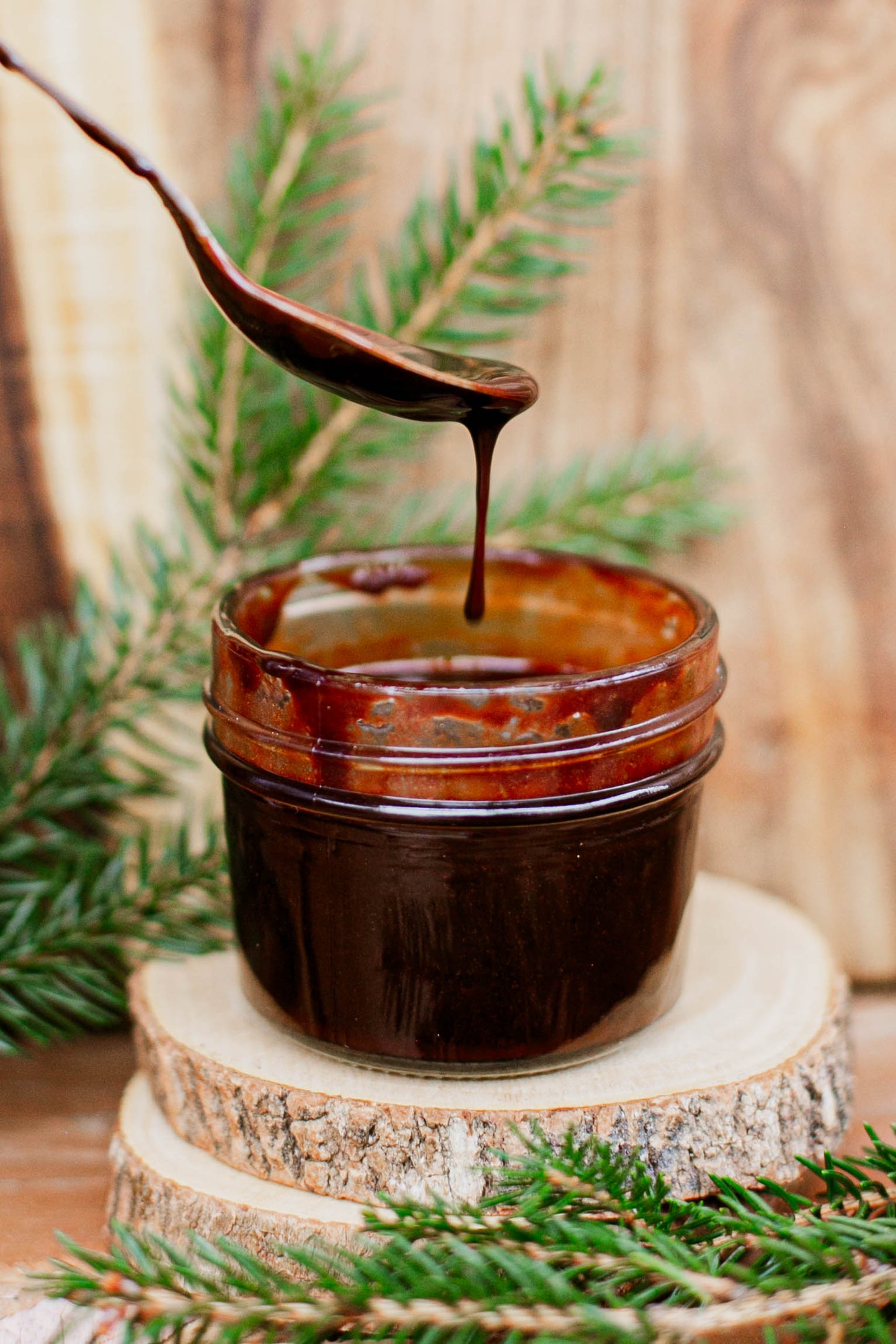 Glass jar with fudge syrup and a spoon drizzling syrup back into the jar on a wooden surface with greenery. Made in the kitchen at Nystrom Orchard in Worthington Minnesota.