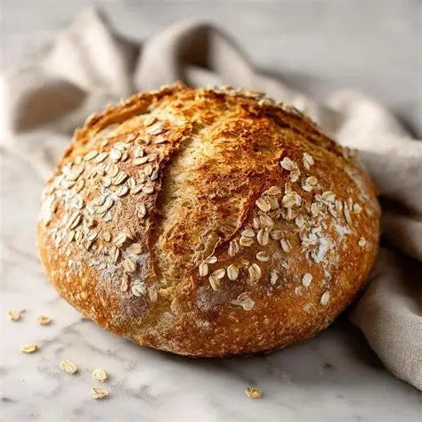 A round artisanal bread loaf topped with rolled oats and a golden-brown crust, resting on a light marble surface with a beige cloth in the background. Made in the kitchen of Nystrom Orchard.