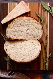 Rustic loaf of sliced parmesan and herb sourdough bread on a wooded cutting board with fresh herbs and a wedge of parmesan beside it. Baked in the kitchen at Nystrom Orchard in Worthington Minnesota.