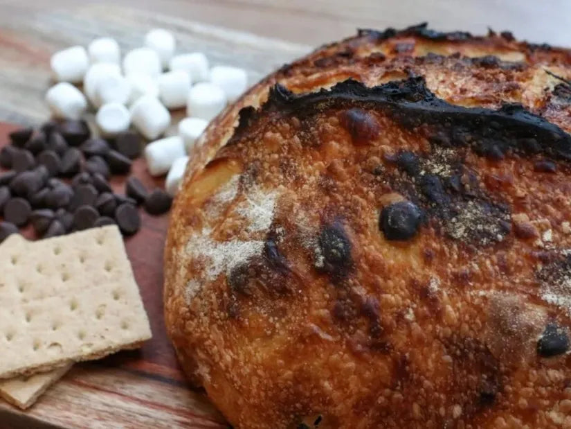 Artisan loaf of sourdough bread with graham crackers, chocolate chips and marshmallows swirled inside on a wooden cutting board. Beside the loaf is chocolate chips mini marshmallows and graham crackers. Baked in the kitchen of Nystrom Orchard in Worthington Minnesota.