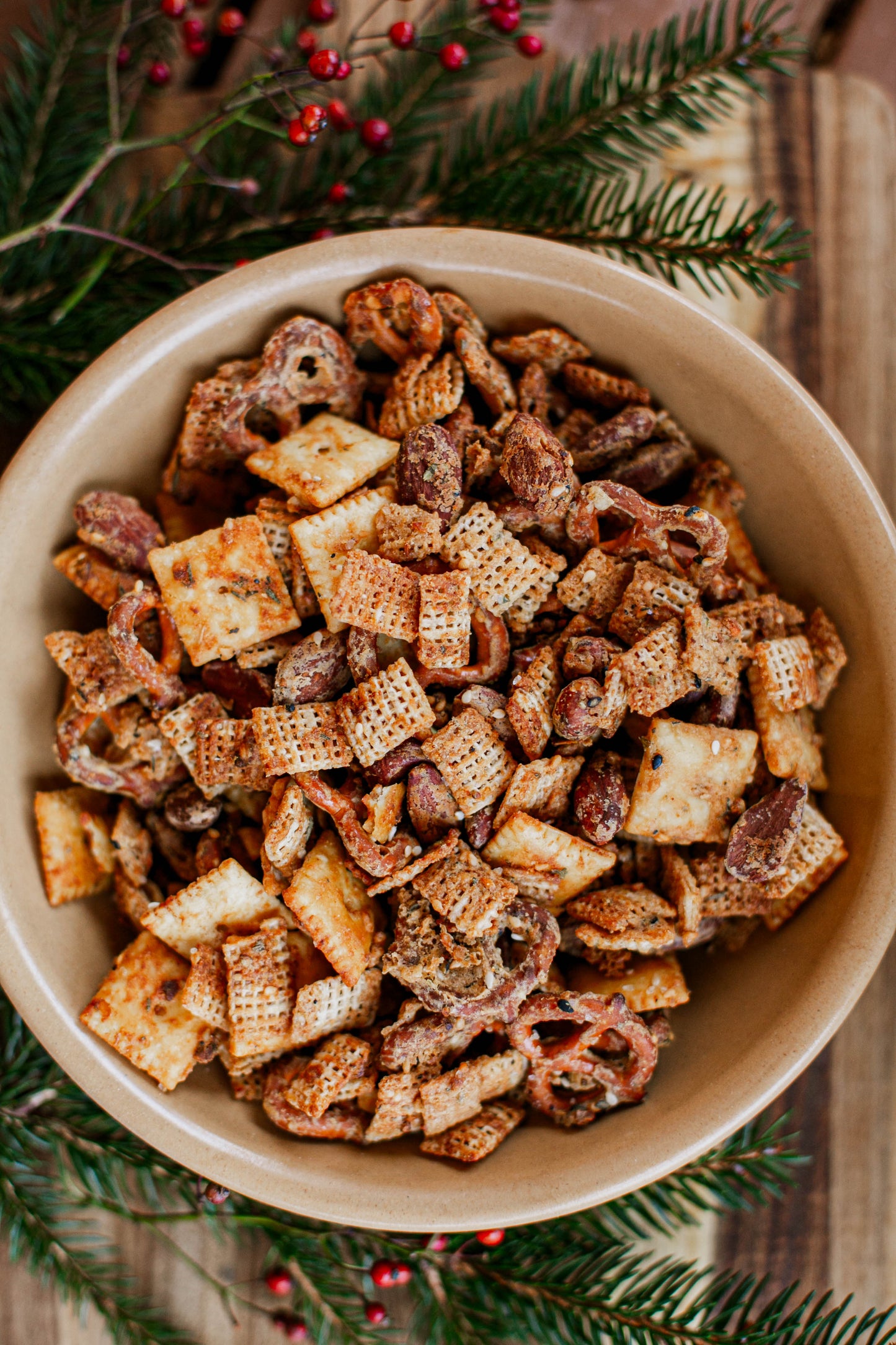 A tan bowl of sourdough snack mix on a wooden board with greenery and red berries. Made in the kitchen at Nystrom Orchard in Worthington Minnesota.