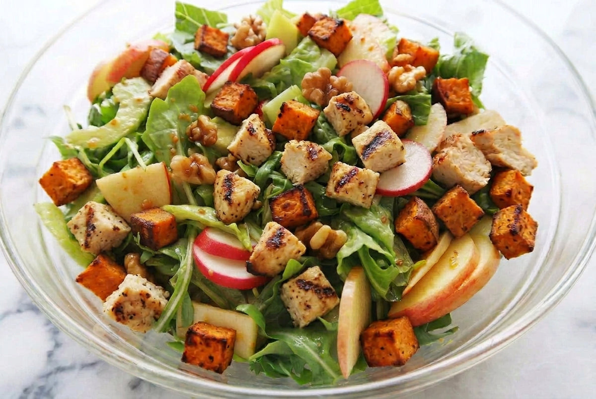 A close-up shot of a fresh salad in a clear glass bowl, featuring mixed greens, grilled chicken breast cubes, caramelized sweet potato cubes, sliced radishes, apple pieces, and walnuts on a light marble surface. Made by Nystrom Orchard in Worthington Minnesota.