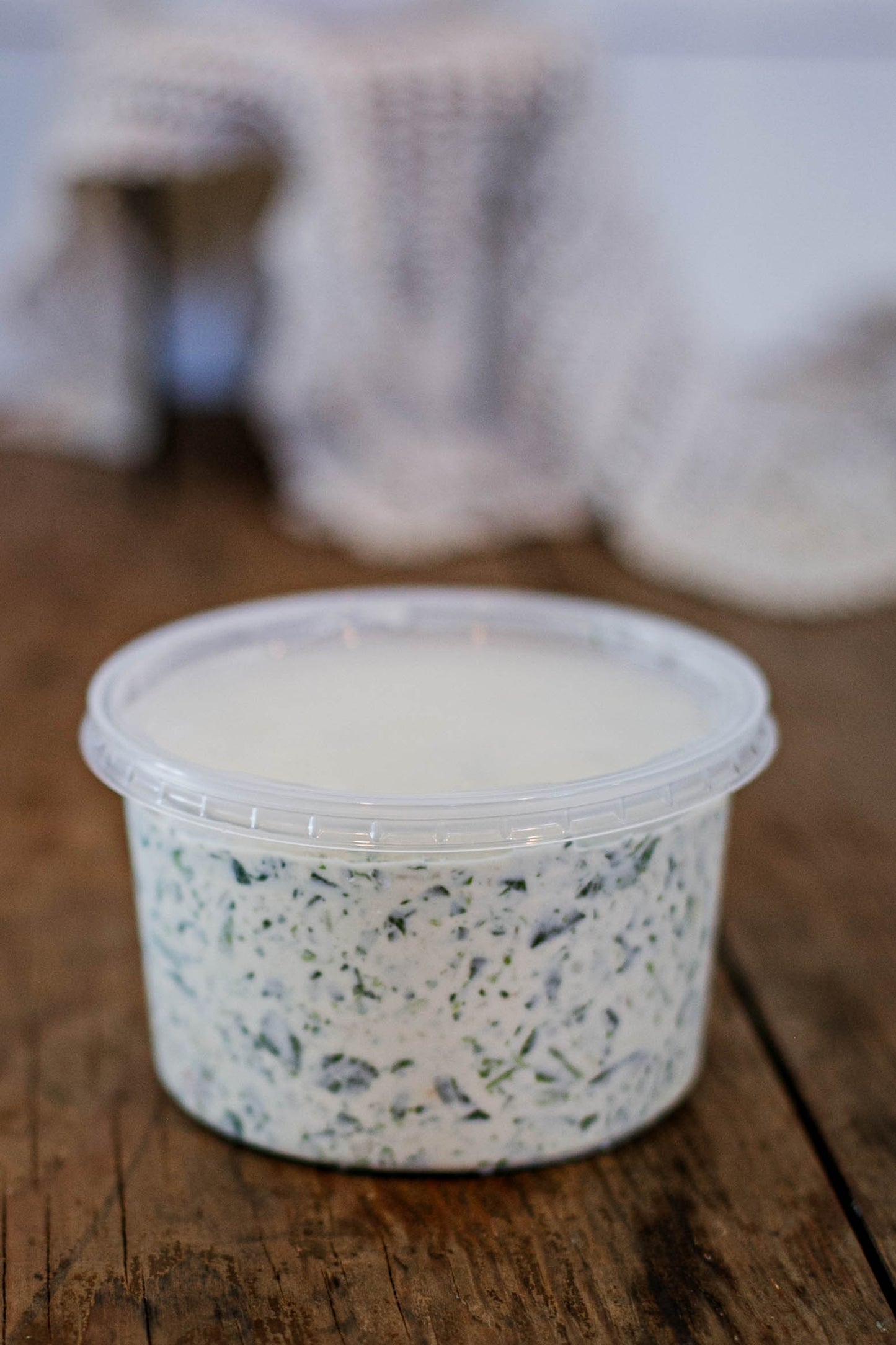 Container of creamy spinach dip in a clear container with a lid on a wooden surface and a blurred background. Made in the kitchen at Nystrom Orchard in Worthington Minnesota.