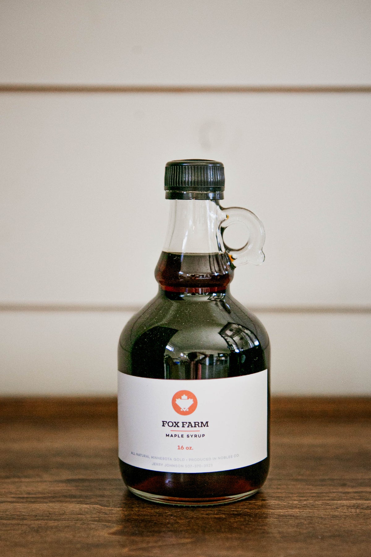 16 oz jug of maple syrup on a wooden table on a white background. Sold by Nystrom Orchard in Worthington Minnesota.