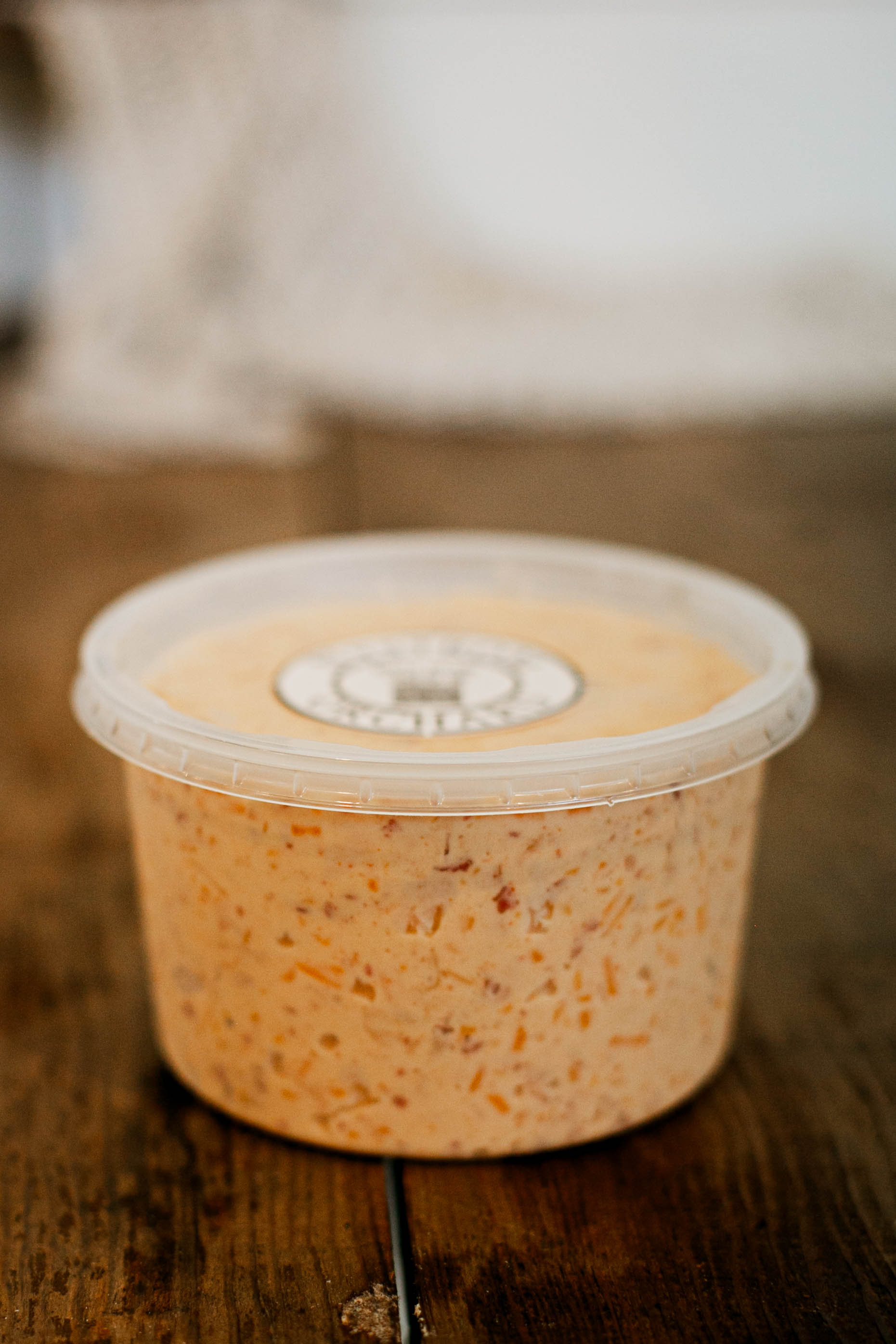 16 oz container of maple bacon cheese dip with a lid on a wooden surface with a blurry background.  Made in the kitchen of Nystrom Orchard in Worthington Minnesota.