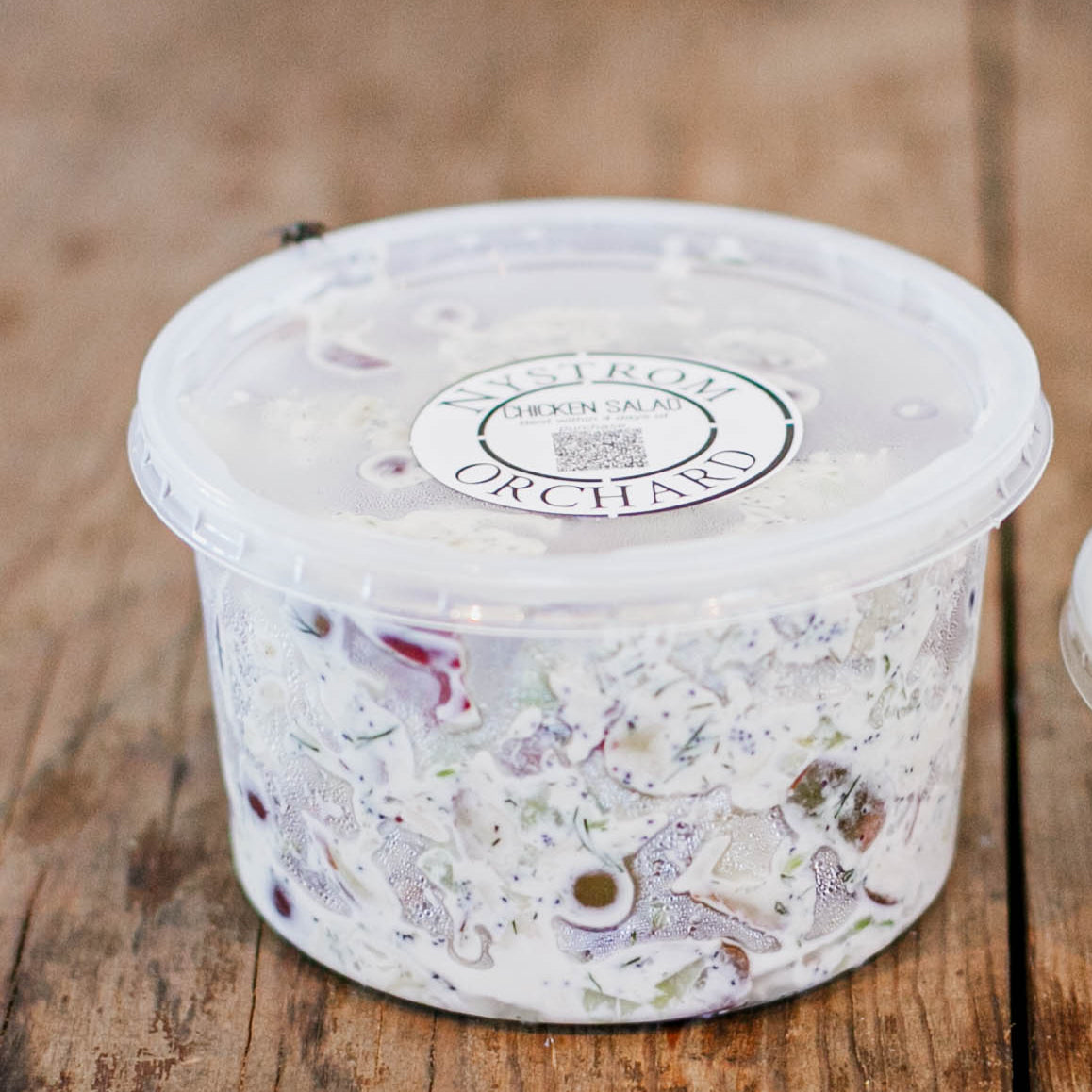 16 oz container of chicken salad with a lid on a wooden surface. Made in the kitchen of Nystrom Orchard in Worthington Minnesota.