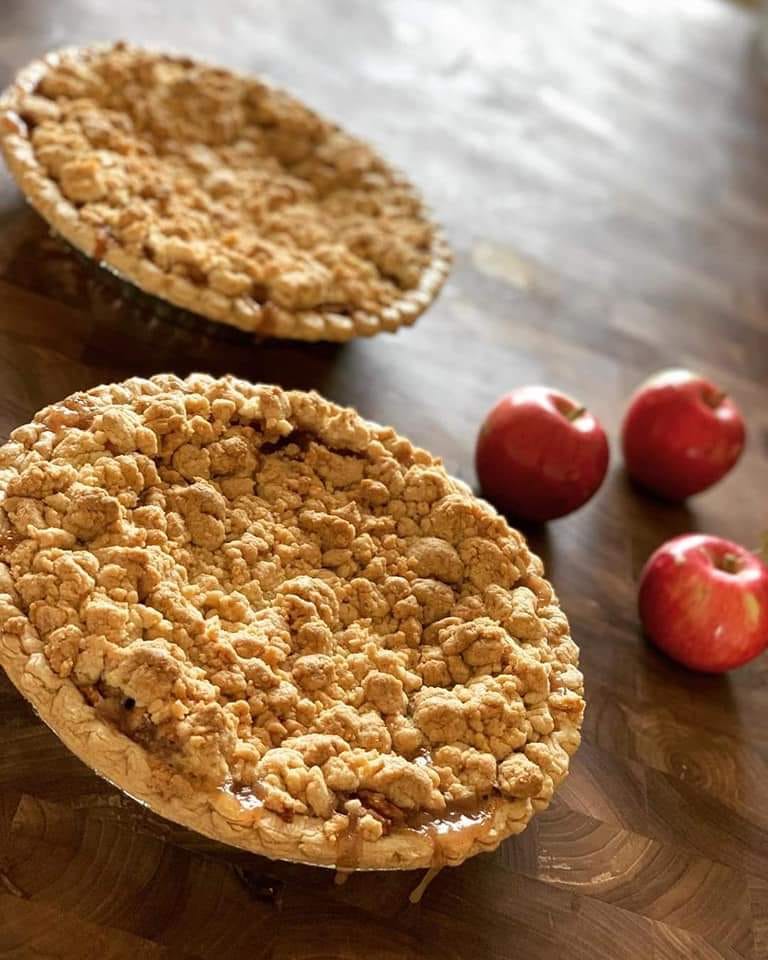 Two baked apple pies on a wooden surface with apples setting beside. Made by Nystrom Orchard in Worthington Minnesota.