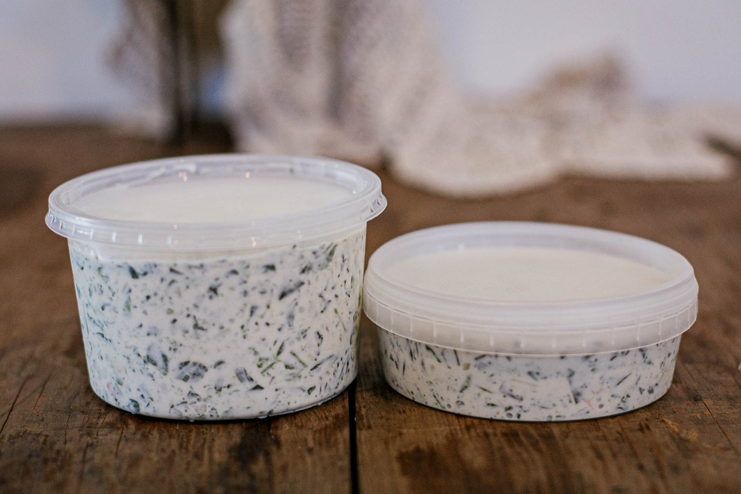 Two containers of creamy spinach dip with lids on a wooden surface with a blurred background. Made in the kitchen of Nystrom Orchard in Worthington Minnesota.