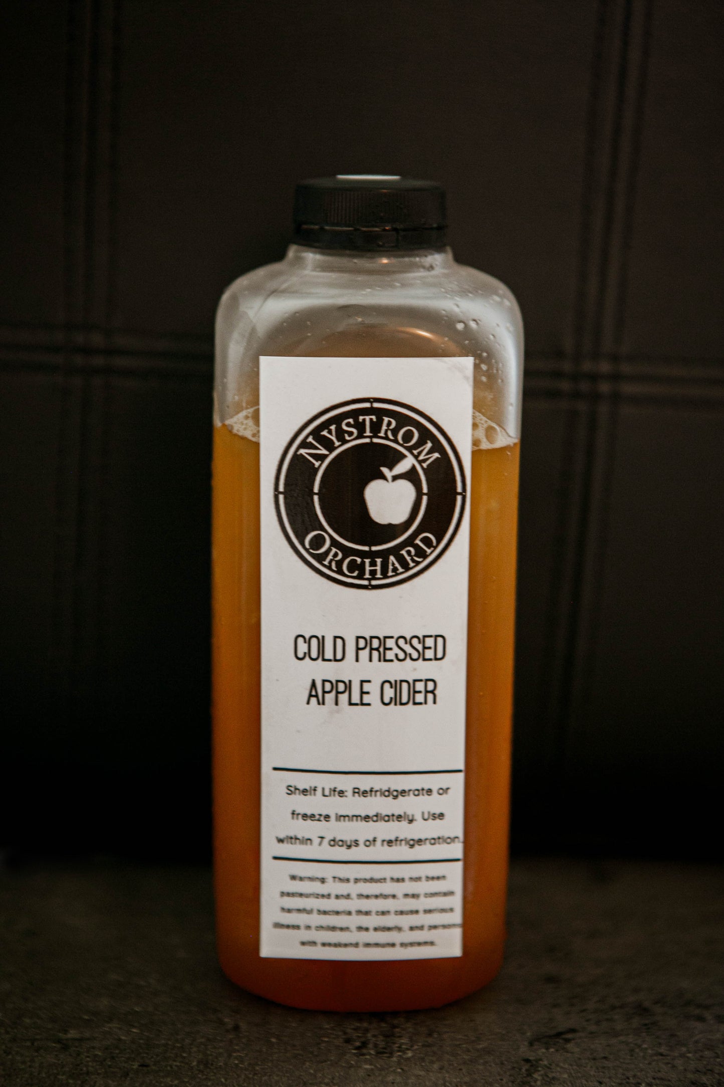 Bottle of cold pressed apple cider with a label on a dark background. Made in the kitchen at Nystrom Orchard in Worthington Minnesota.