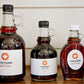 Three bottles of maple syrup arranged from largest to smallest on a wooden surface, with labels showing the brand 'Fox Farm'. Sold by Nystrom Orchard in Worthington Minnesota.