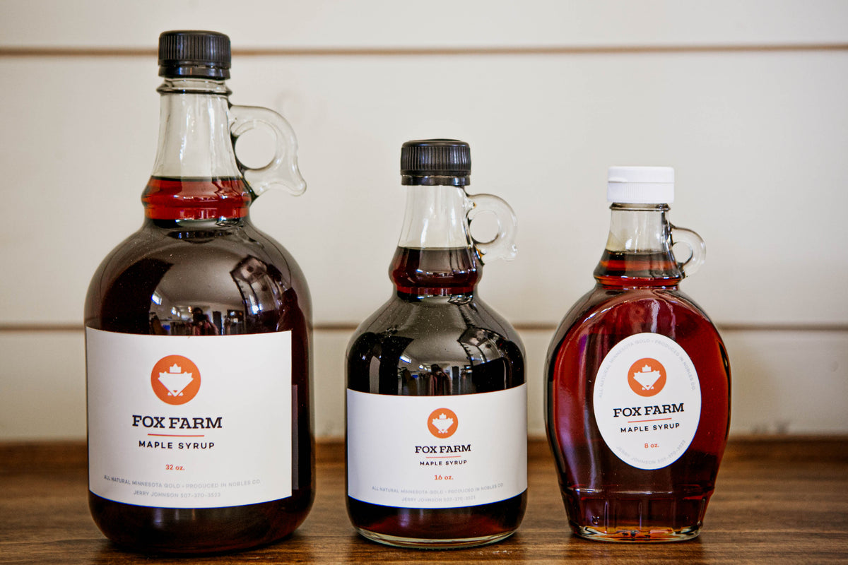 Three bottles of maple syrup arranged from largest to smallest on a wooden surface, with labels showing the brand 'Fox Farm'. Sold by Nystrom Orchard in Worthington Minnesota.