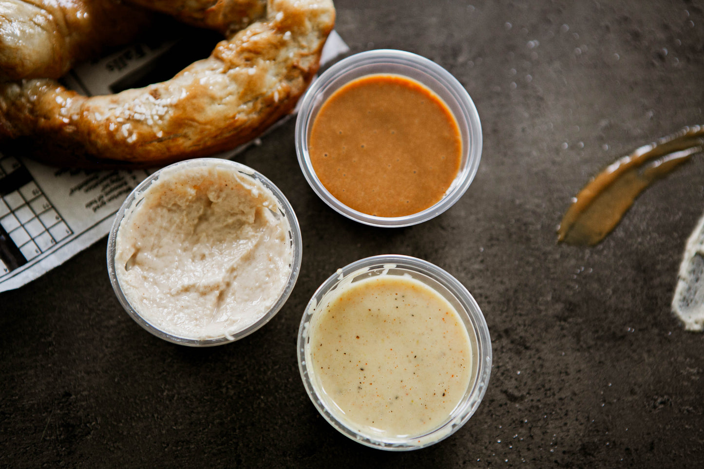 3- 2 oz containers of house made dips with a partial pretzel beside on a dark background. Made in the kitchen of Nystrom Orchard in Worthington Minnesota. 