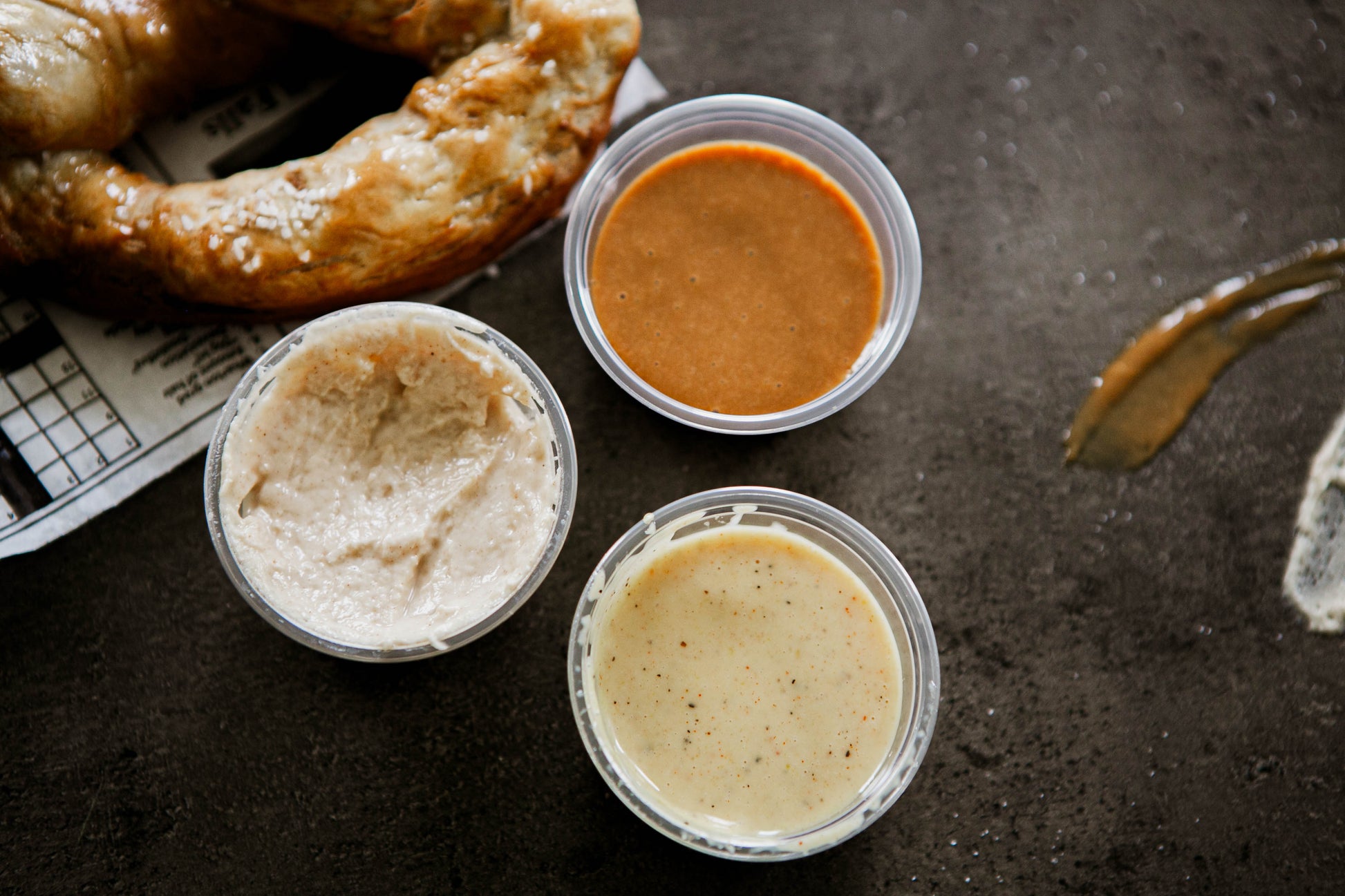 3- 2 oz containers of house made dips with a partial pretzel beside on a dark background. Made in the kitchen of Nystrom Orchard in Worthington Minnesota. 