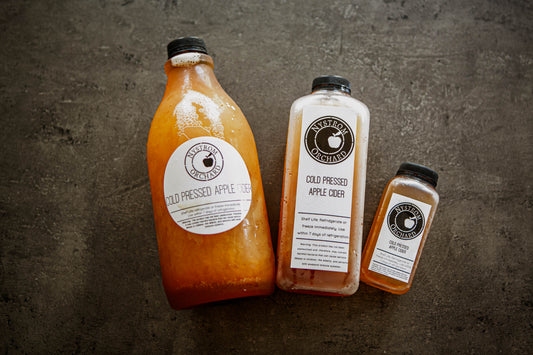 Three bottles of cold pressed cider on a textured surface, with varying sizes and labels indicating different types of cider. Made in the kitchen of Nystrom in Worthington Minnesota.