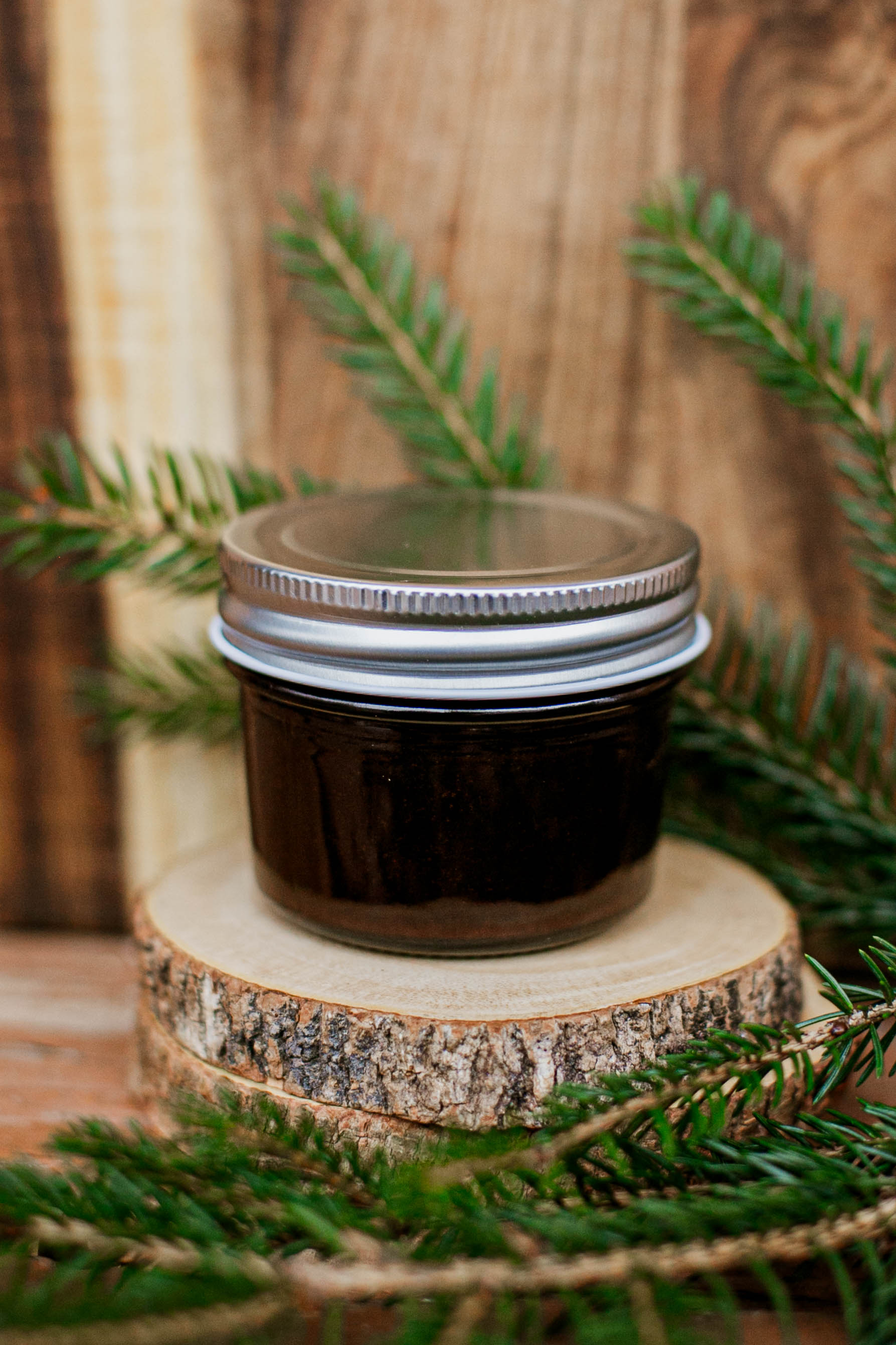 Jar of chocolate fudge sauce with a metal lid on a wooden stand with greenery. Made in the kitchen at Nystrom Orchard in Worthington Minnesota.