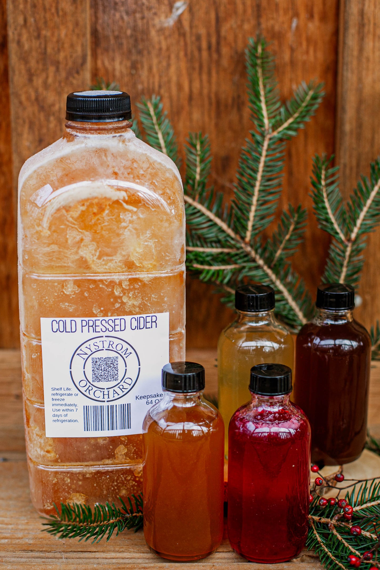 64 oz cold pressed cider with 4 bottles of cider syrup on a wooden surface with greenery and red berries. Made in the kitchen at Nystrom Orchard in Worthington Minnesota.