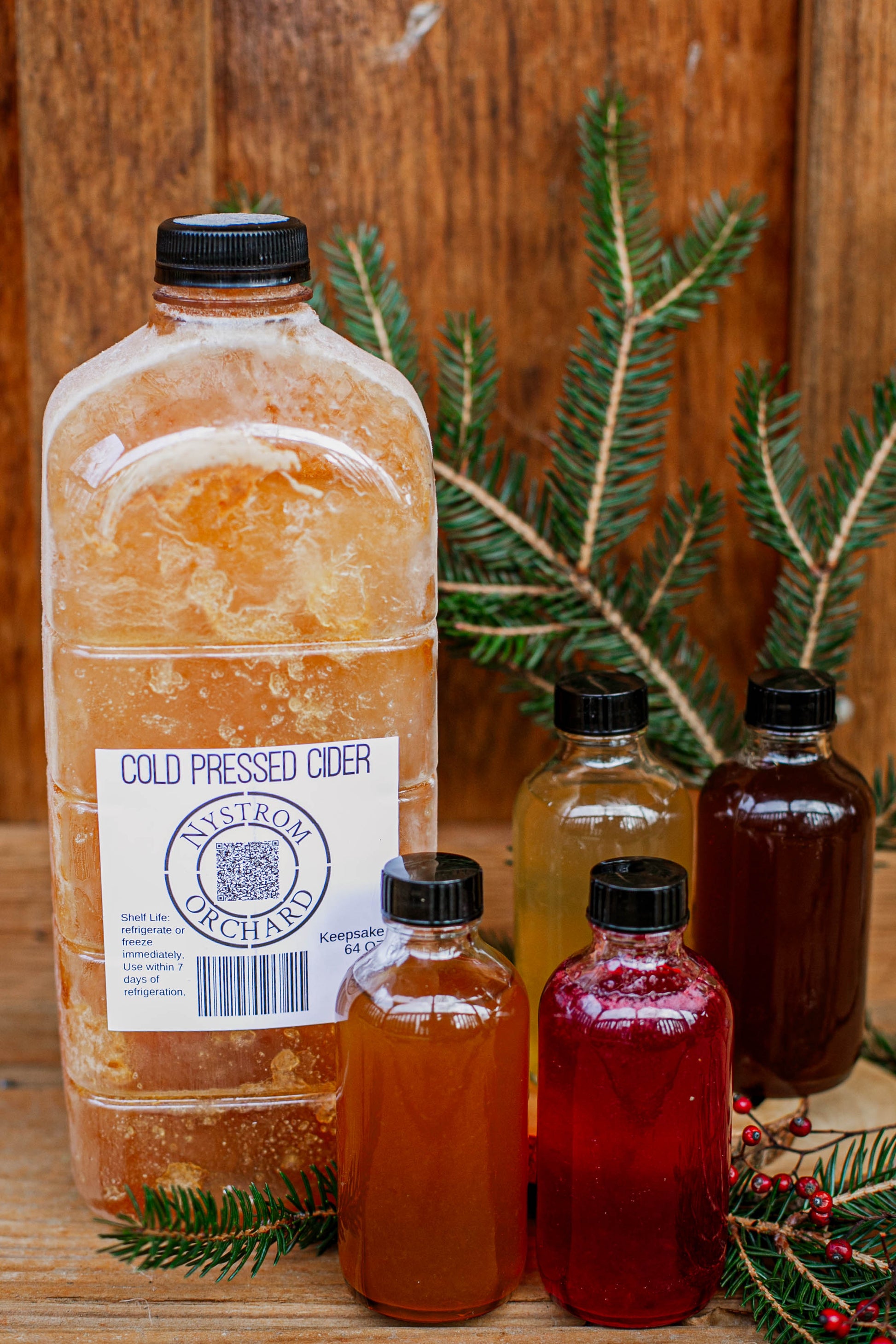64 oz cold pressed cider with 4 bottles of cider syrup on a wooden surface with greenery and red berries. Made in the kitchen at Nystrom Orchard in Worthington Minnesota.