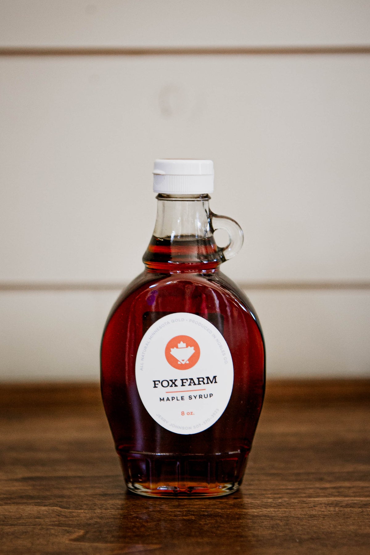 8 oz jug of maple syrup on a wooden surface with a white background. Sold by Nystrom Orchard in Worthington Minnesota.