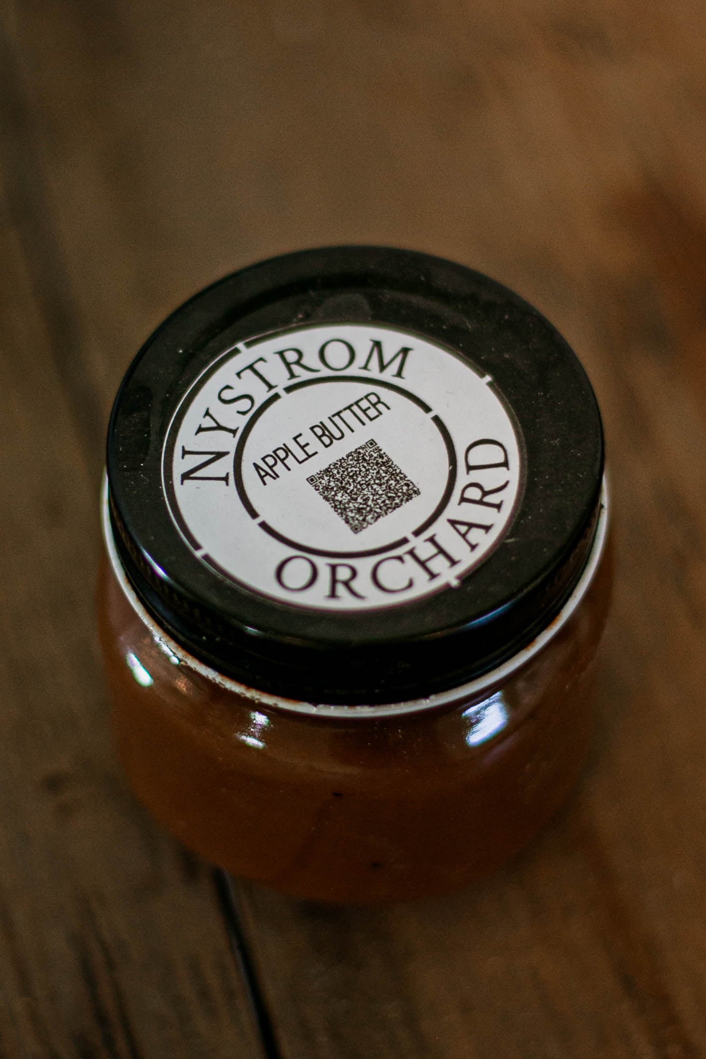 Jar of Nystrom Orchard apple butter with a black lid on a wooden surface. Made in the kitchen of Nystrom Orchard in Worthington Minnesota.