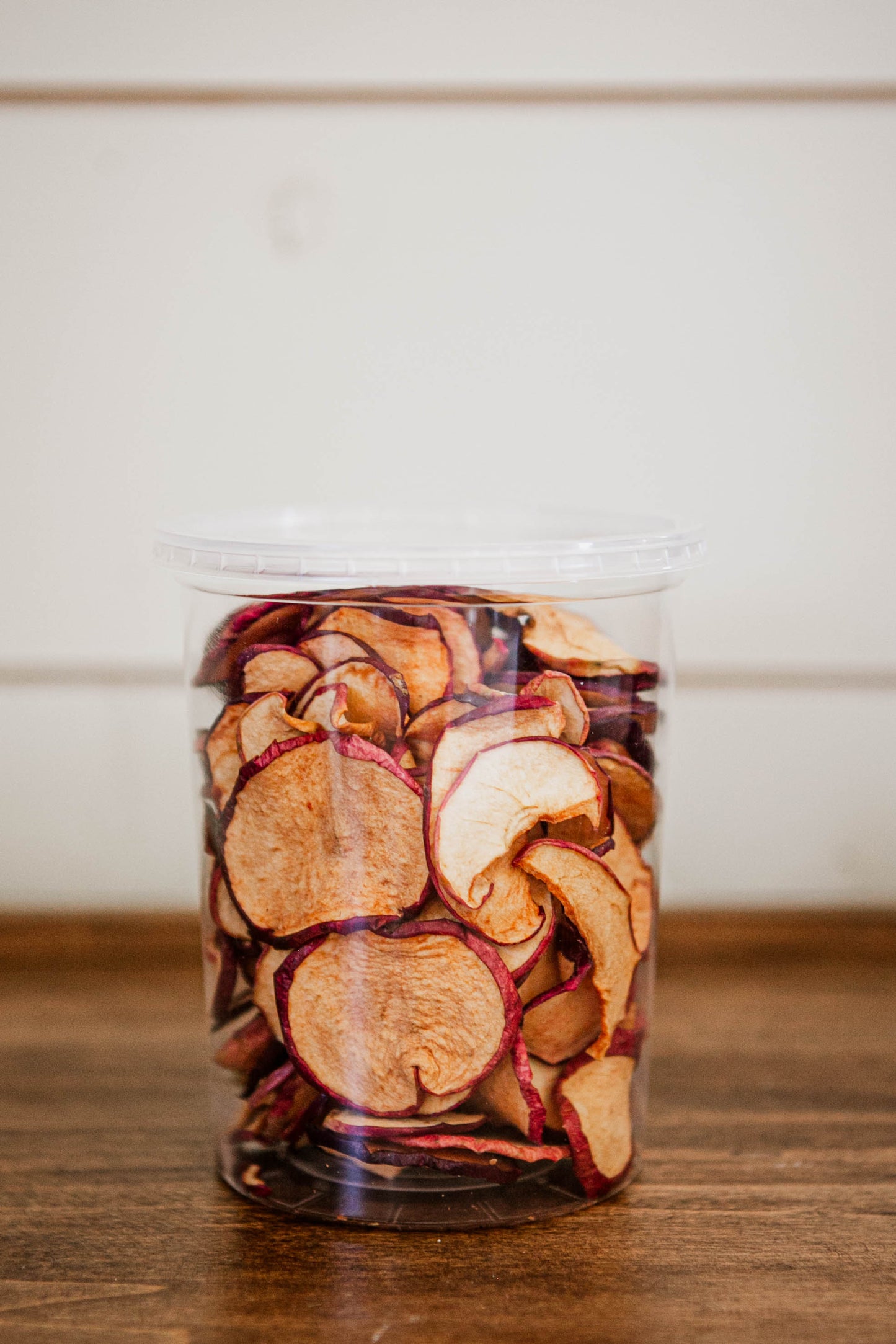 Dried apple chips in a clear 16 oz container on a wooden surface with a white background. Dried in the kitchen of Nystrom Orchard in Worthington Minnesota.