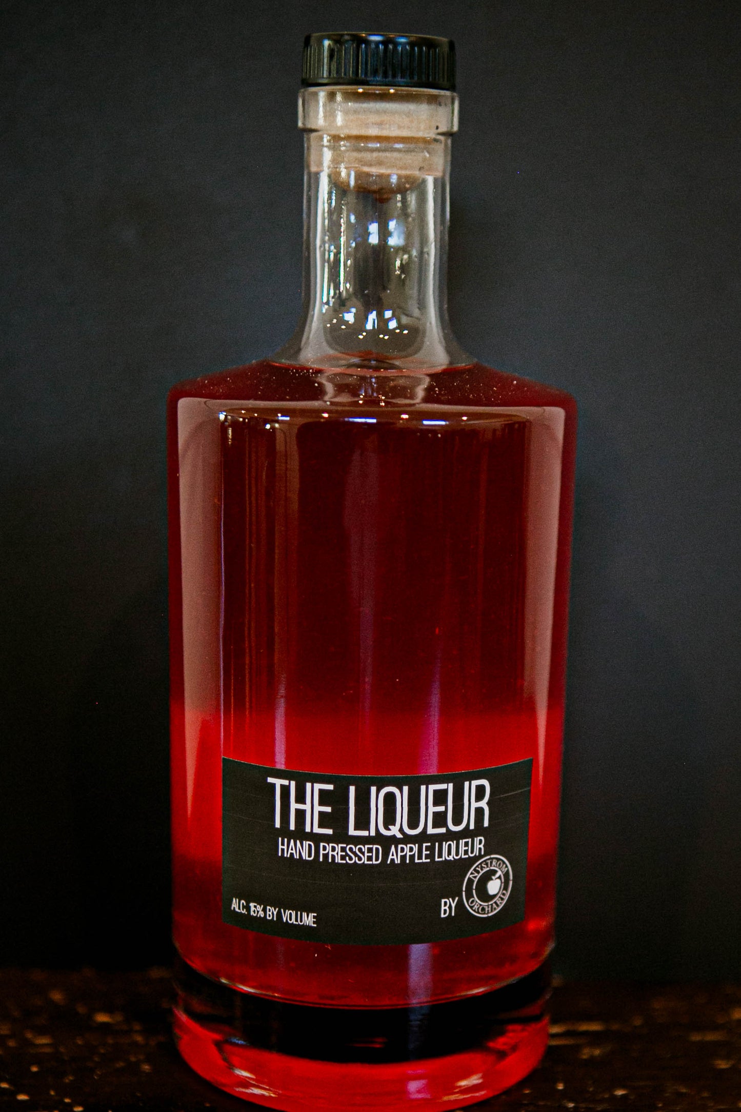 Bottle of pink liqueur in a glass bottle with a black lid on a black background. Hand crafted in the kitchen at Nystrom Orchard in Worthington Minnesota.