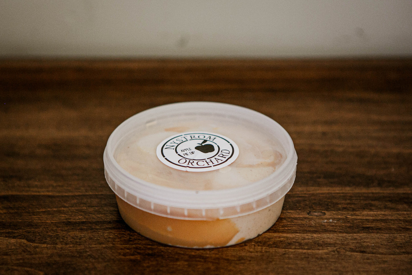 Apple pie dip in a clear plastic container topped with caramel on a wooden surface. Made in the kitchen of Nystrom Orchard in Worthington Minnesota.