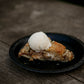A slice of pie topped with a scoop of ice cream on a black plate, placed on a wooden table. Made in the kitchen of Nystrom Orchard in Worthington Minnesota.