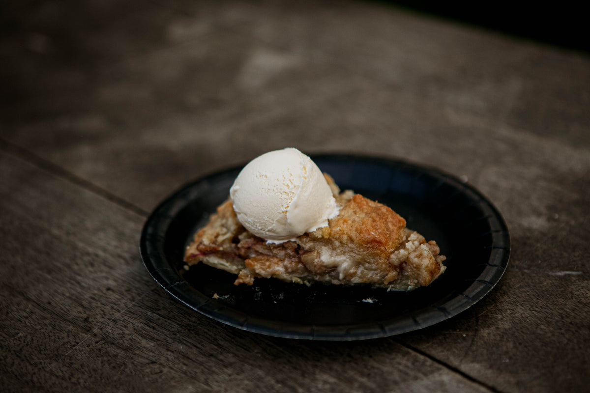 A slice of pie topped with a scoop of ice cream on a black plate, placed on a wooden table. Made in the kitchen of Nystrom Orchard in Worthington Minnesota.