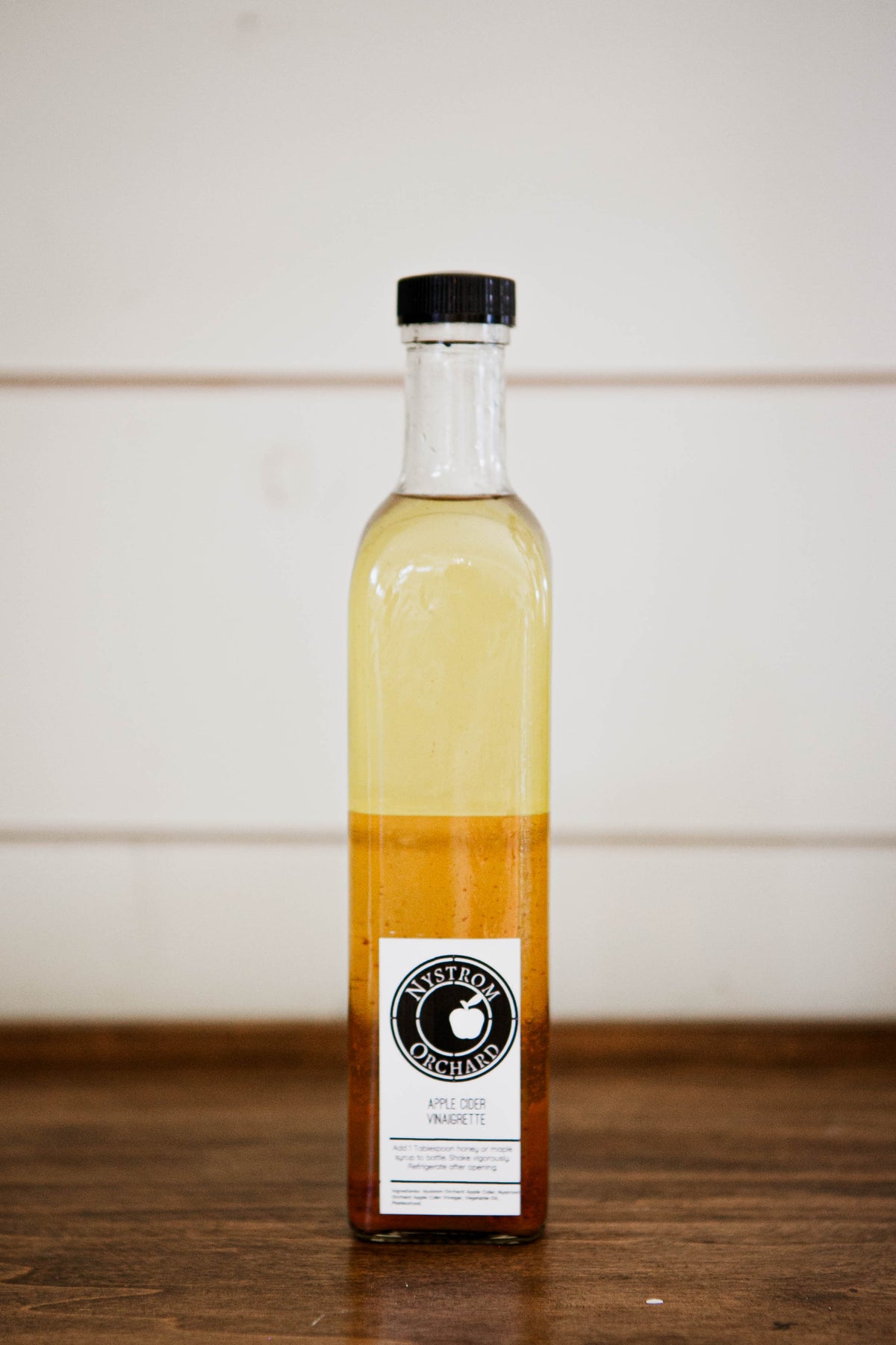 Bottle of apple cider vinaigrette made with vinegar from Nystrom Orchard on a wooden surface with a white background. Made in the kitchen at Nystrom Orchard in Worthington Minnesota.
