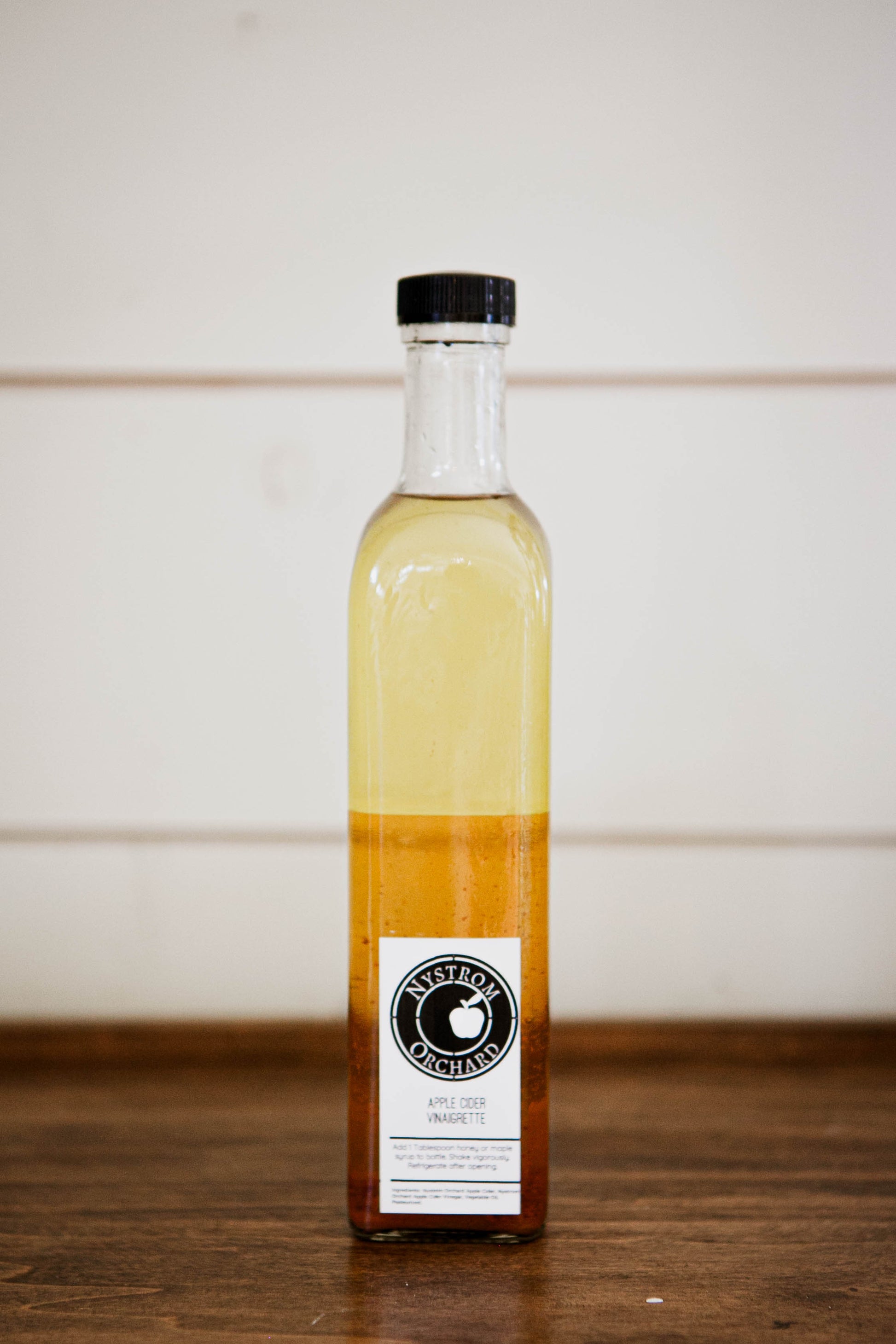 Bottle of apple cider vinaigrette made with vinegar from Nystrom Orchard on a wooden surface with a white background. Made in the kitchen at Nystrom Orchard in Worthington Minnesota.