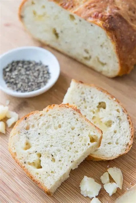 A rustic loaf of sourdough bread sliced on a cutting board with a loaf in the background. There is cheese and black pepper beside the slices of bread. Baked in the kitchen of Nystrom Orchard in Worthington Minnesota. 