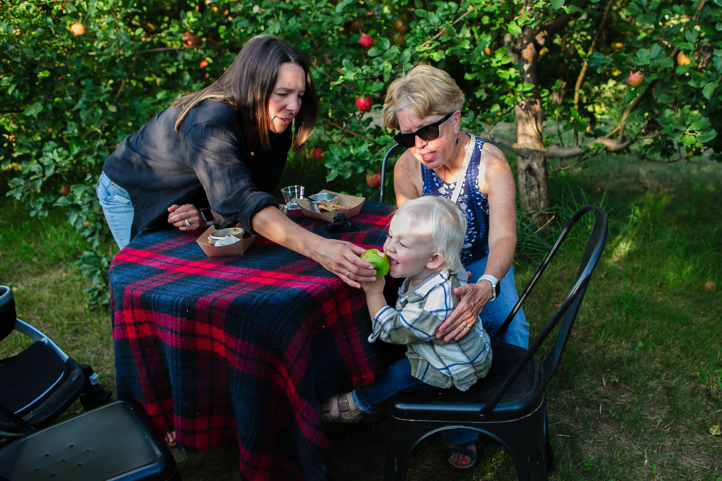 Baby eating an apple with two ladies sitting at a table in an apple orchard. Event hosted by Nystrom Orchard in Worthington Minnesota.