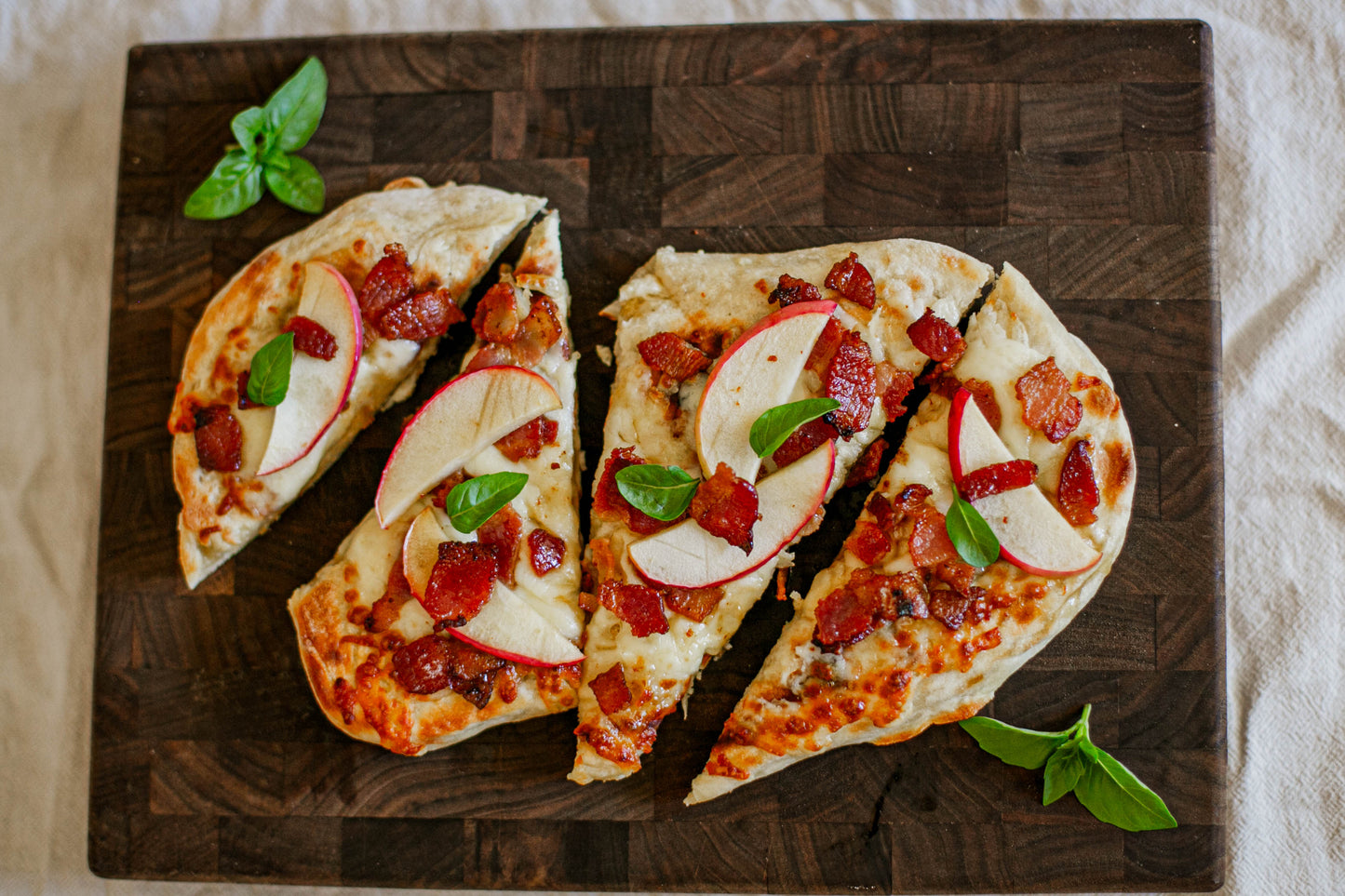 Sliced flatbread pizza with bacon cheddar and apple and basil on a dark cutting board on a white surface. Baked in the kitchen of Nystrom Orchard in Worthington Minnesota.