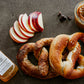 Two pretzels with cold pressed cider, sliced apples and a jar of caramel sauce on a dark surface. Made in the kitchen of Nystrom Orchard in Worthington Minnesota.