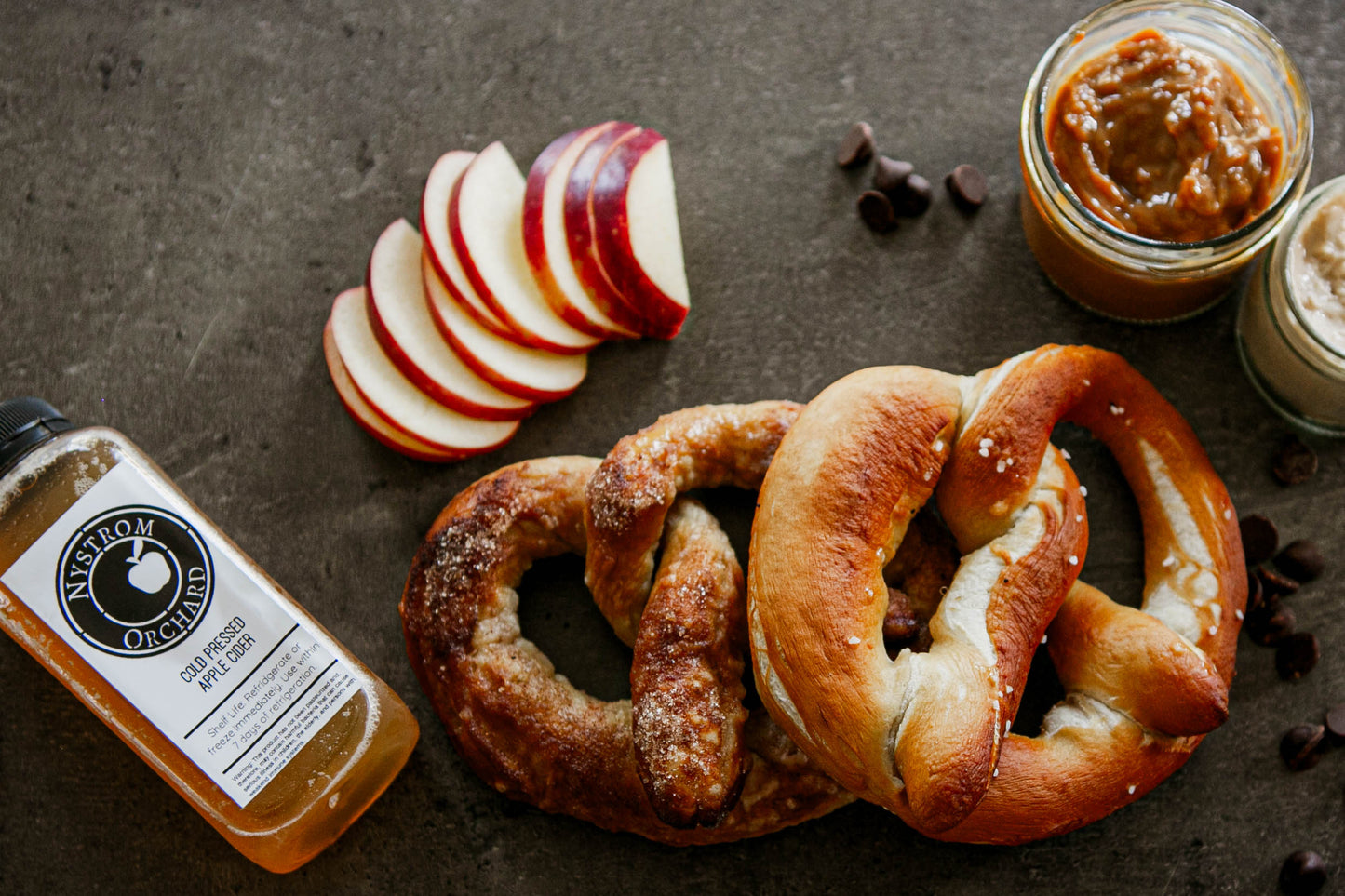Two pretzels with cold pressed cider, sliced apples and a jar of caramel sauce on a dark surface. Made in the kitchen of Nystrom Orchard in Worthington Minnesota.