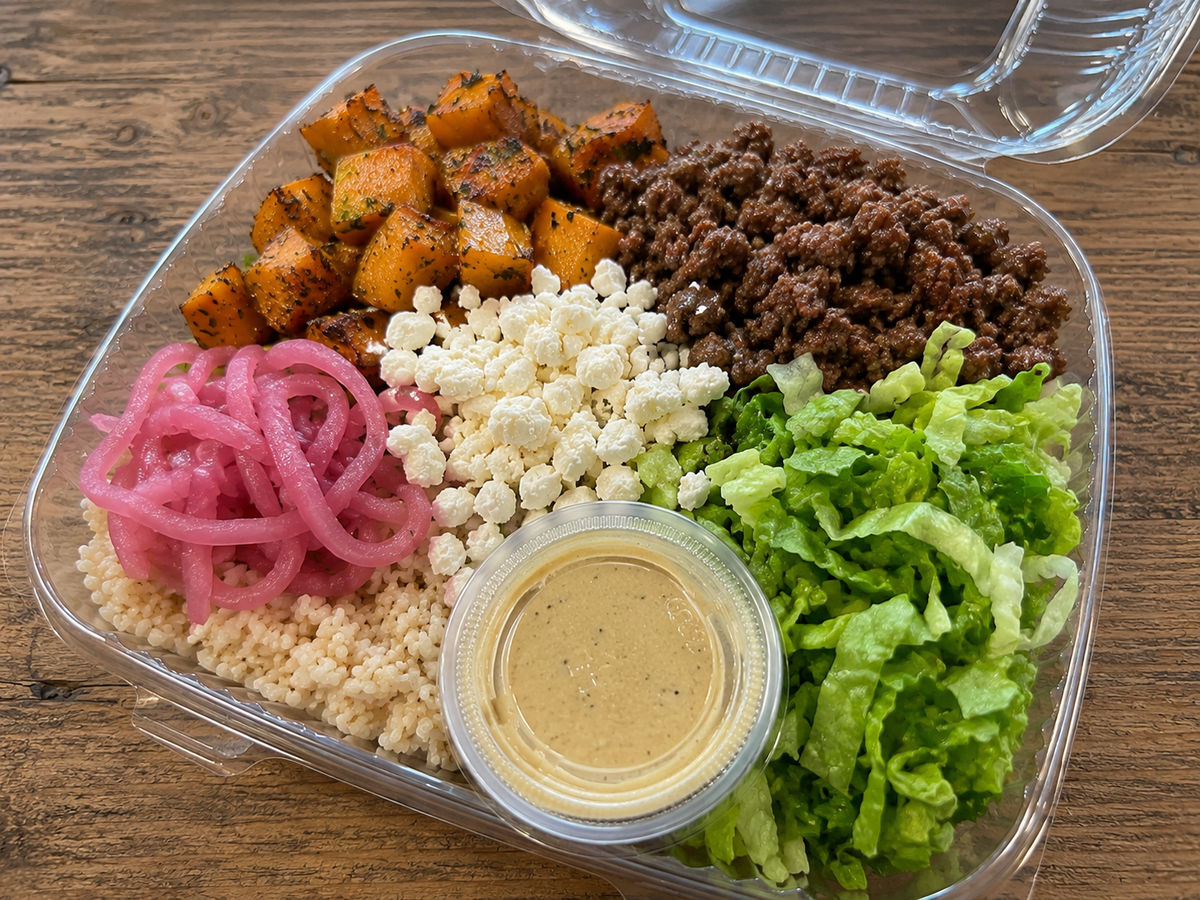 A clear plastic deli container filled with a prepared meal on a wooden table, featuring roasted squash cubes, ground beef, pickled onions, wild rice, feta, lettuce, and a small cup of honey mustard dressing. Make in the kitchen of Nystrom Orchard in Worthington Minnesota.