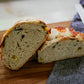 Rustic loaf of basil mozerella sourdough bread cut in half so you can see the crumb. It's on a wooden cutting board with a bread knife and black and white striped towel. Baked in the kitchen at Nystrom Orchard in Worthington Minnesota.