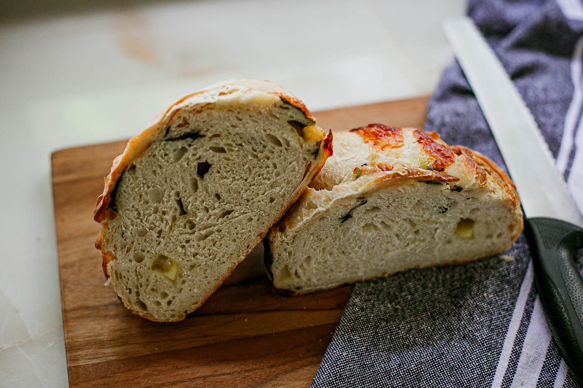 Rustic loaf of basil mozerella sourdough bread cut in half so you can see the crumb. It's on a wooden cutting board with a bread knife and black and white striped towel. Baked in the kitchen at Nystrom Orchard in Worthington Minnesota.