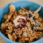 Blue glass bowl full of homemade sourdough granola that includes dried appleson a wooden surface. Made in the kitchen of Nystrom Orchard in Worthington Minnesota.