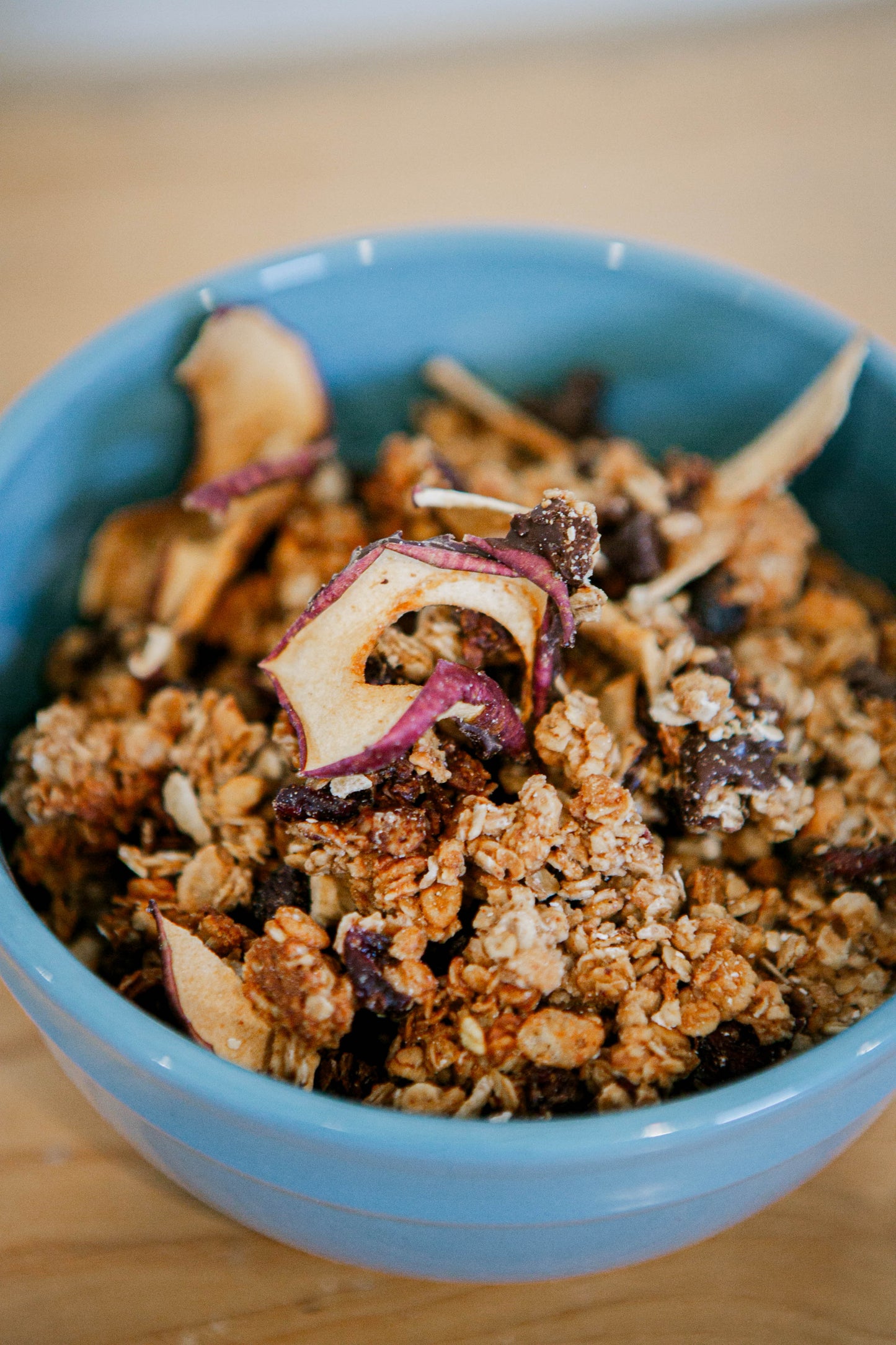 Blue glass bowl full of homemade sourdough granola that includes dried appleson a wooden surface. Made in the kitchen of Nystrom Orchard in Worthington Minnesota.