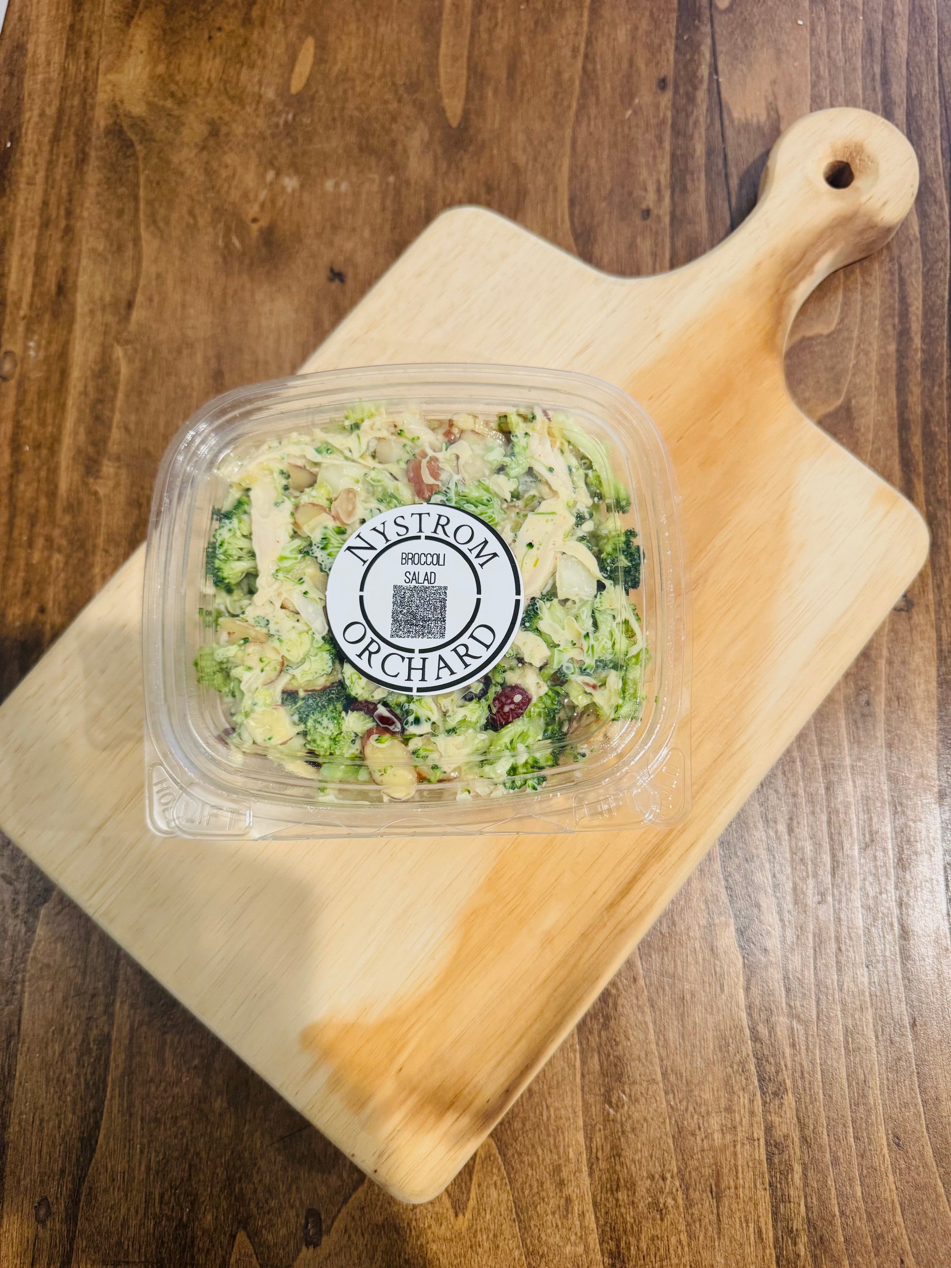 A clear plastic 8 oz container of broccoli salad with chicken on a wooden cutting board. Made in the kitchen of Nystrom Orchard in Worthington Minnesota.