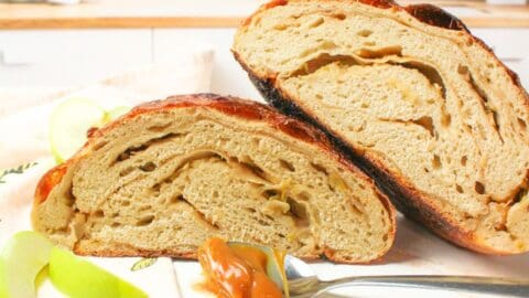 Rustic loaf of caramel apple sourdough bread cut in half on a white plate with some caramel on a spoon and two slices of apples. Baked in the kitchen at Nystrom Orchard in Worthington Minnesota.