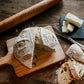 A rustic loaf of jalapeno cheddar sourdough on a cutting board with a couple of slices laying beside on a wooden table. Beside the bread is a rolling pin and a slate coaster with butter and a butter knife.  Baked in the kitchen of Nystrom Orchard in Worthington Minnesota.