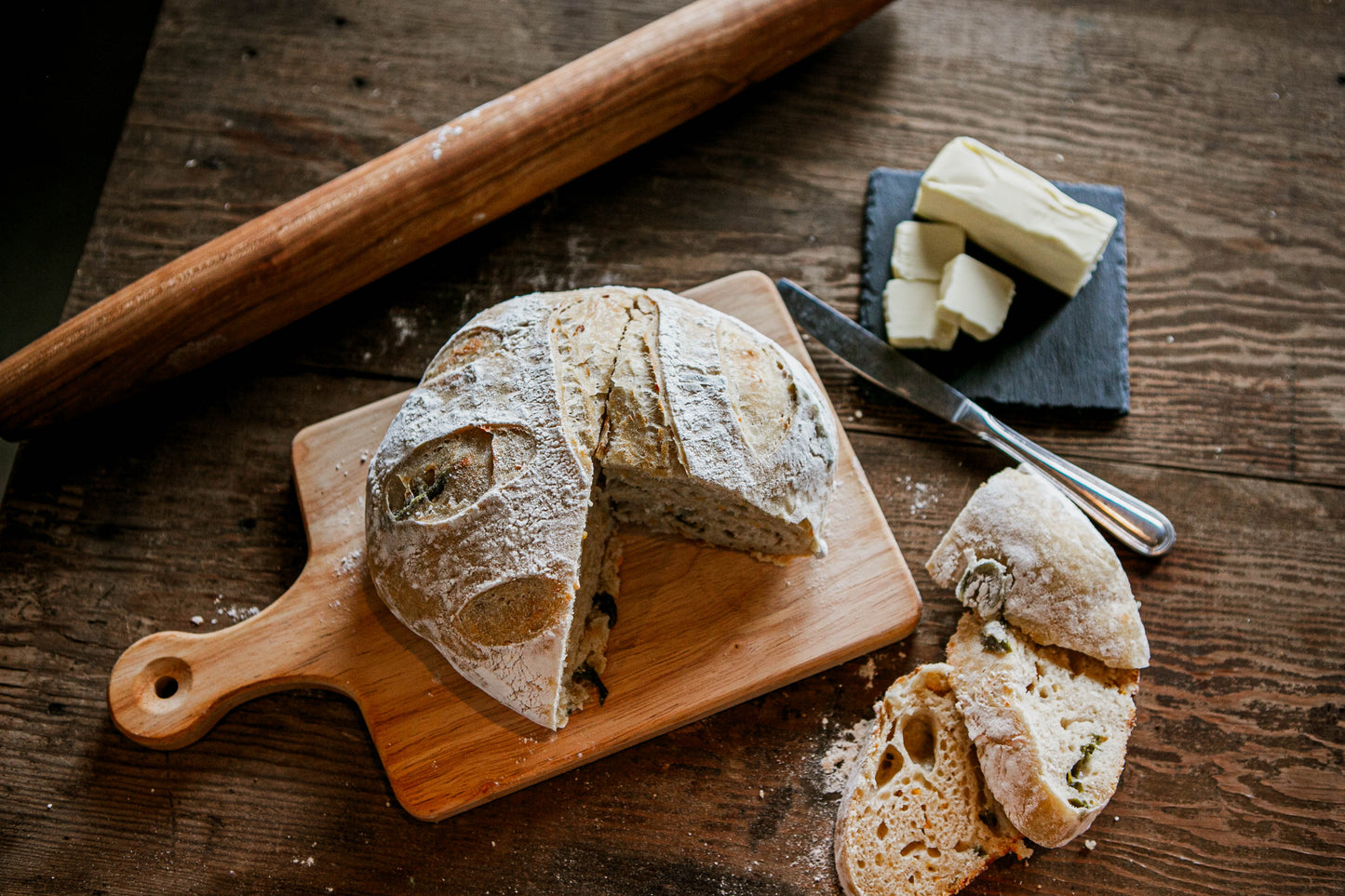 A rustic loaf of jalapeno cheddar sourdough on a cutting board with a couple of slices laying beside on a wooden table. Beside the bread is a rolling pin and a slate coaster with butter and a butter knife.  Baked in the kitchen of Nystrom Orchard in Worthington Minnesota.