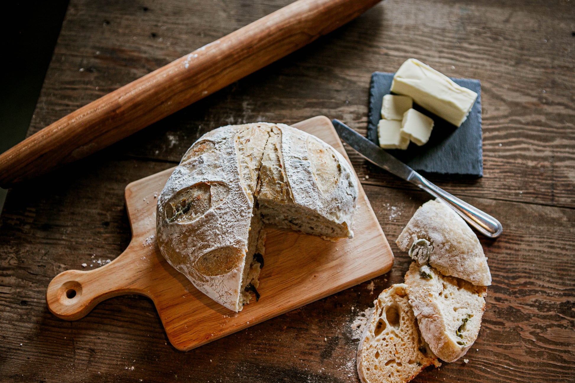A rustic loaf of jalapeno cheddar sourdough on a cutting board with a couple of slices laying beside on a wooden table. Beside the bread is a rolling pin and a slate coaster with butter and a butter knife.  Baked in the kitchen of Nystrom Orchard in Worthington Minnesota.