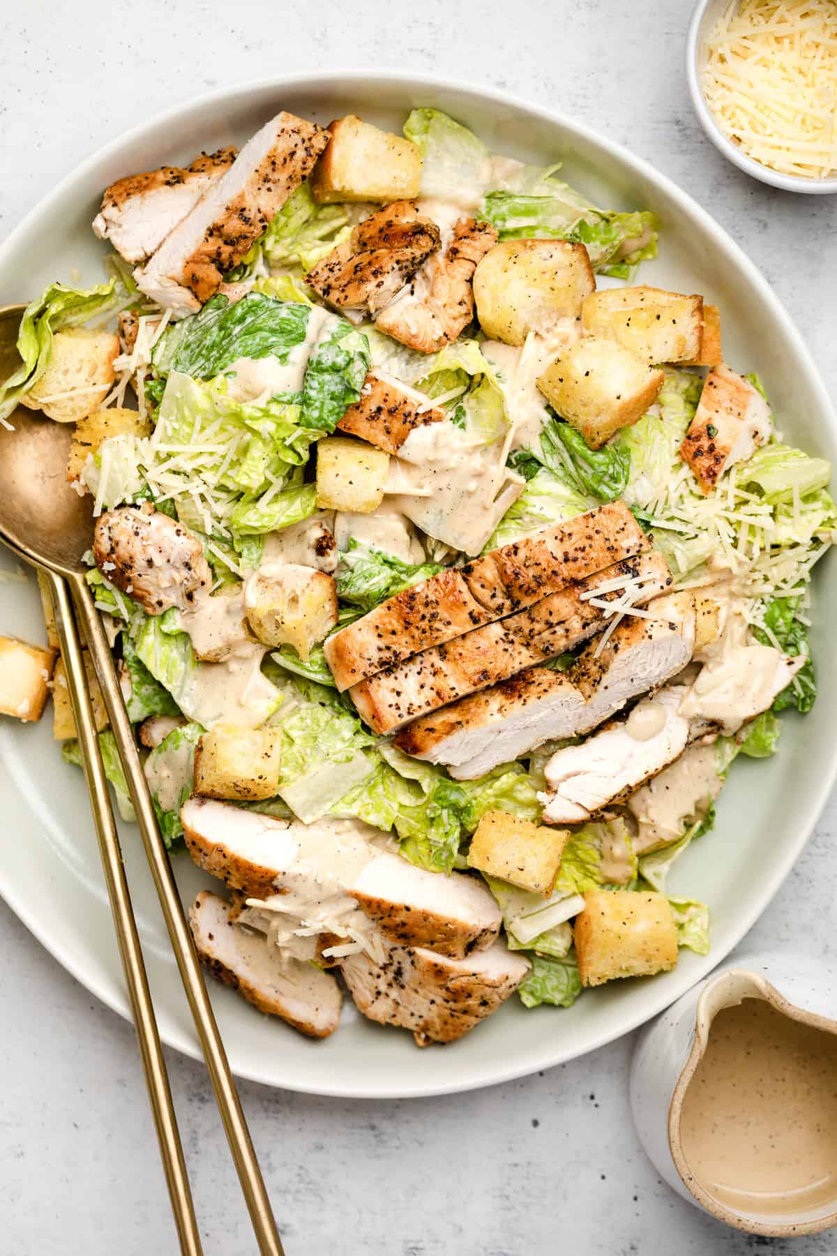 Caesar salad with grilled chicken, croutons, and parmesan cheese on a white plate with gold spoons and dressing container beside. Made in the kitchen at Nystrom Orchard in Worthington Minnesota.